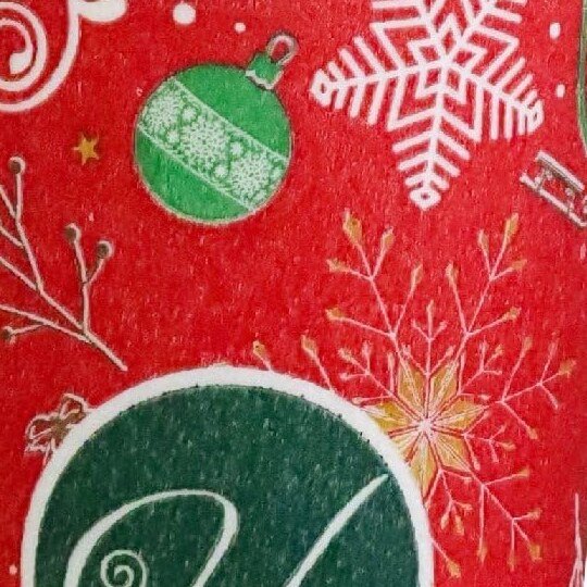 Acercamiento a la superficie afelpada del vaso navideño: el monograma H verde y copos dorados sobre rojo con textura de terciopelo